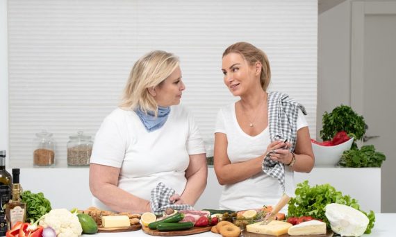 Two women in a kitchen preparing fresh vegetables and cheese with olive oil and spices.